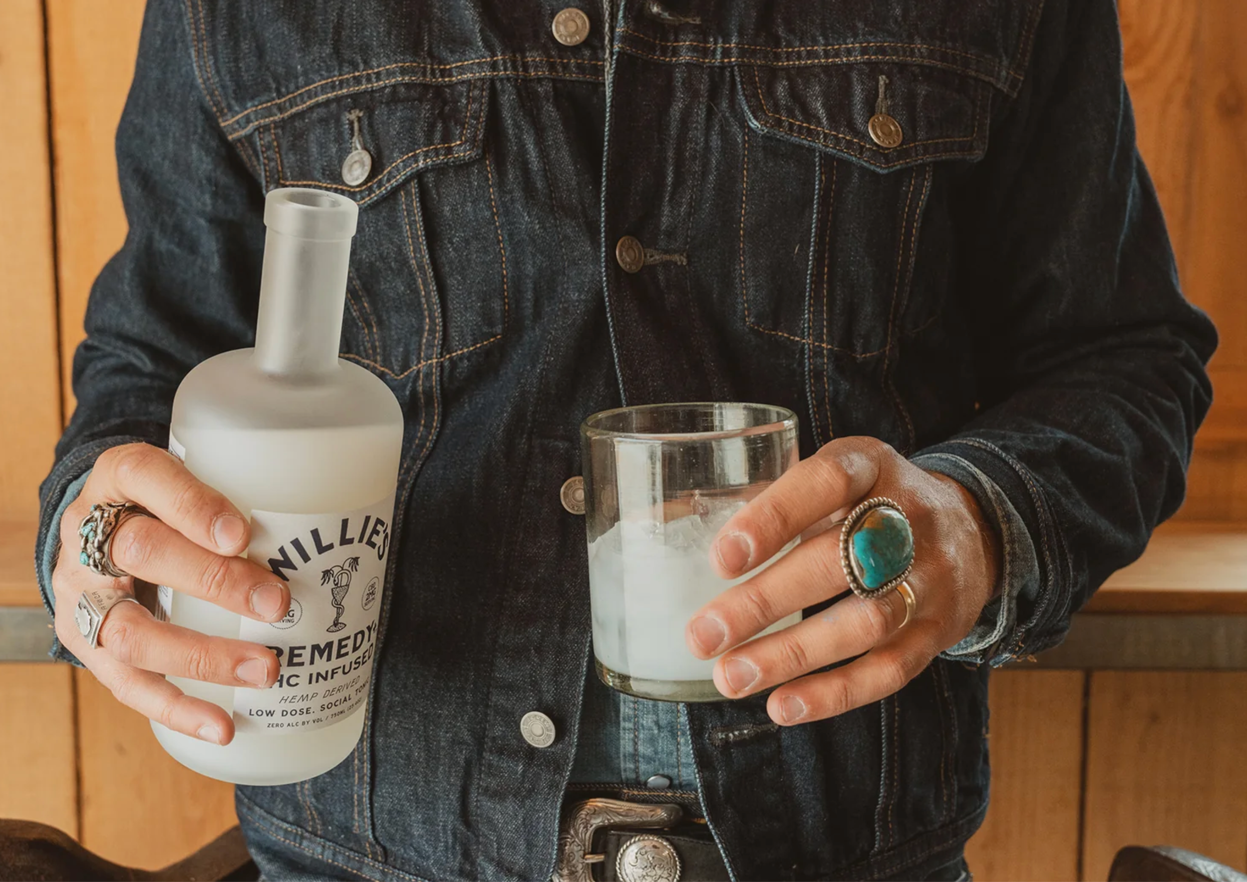 Person holding a bottle and a glass with a drink, wearing a denim jacket.