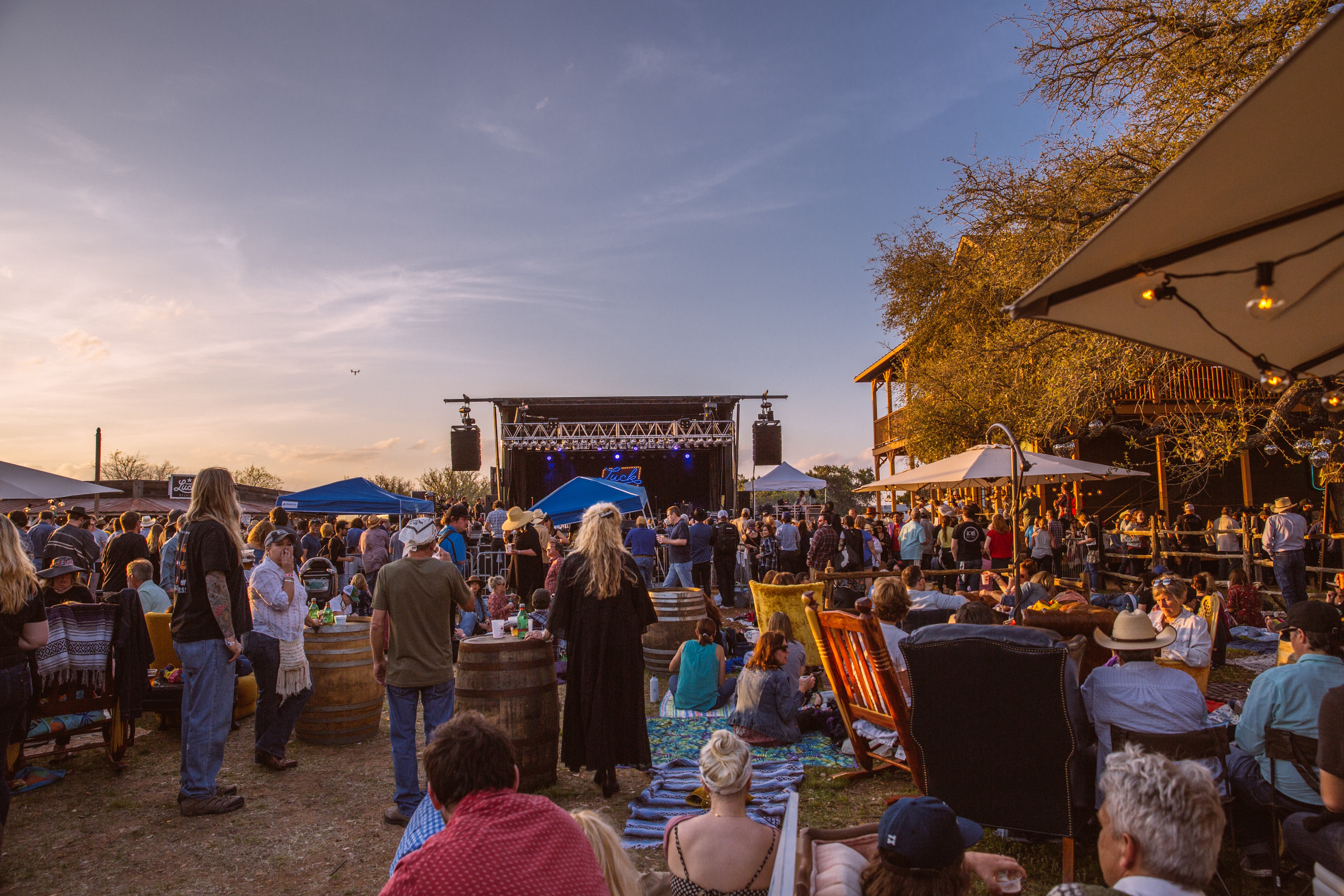 Outdoor event with people gathered around tables and a stage under a blue sky.