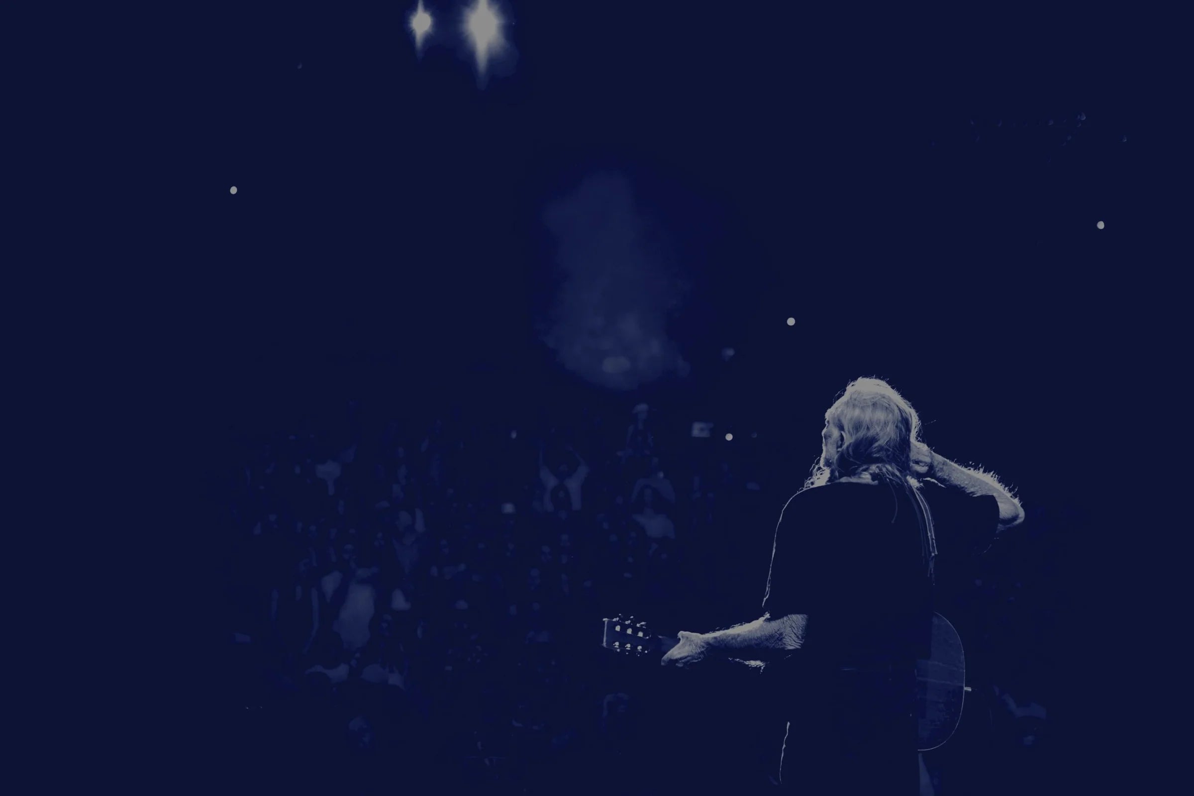 Willie Nelson playing guitar on stage with a dark background