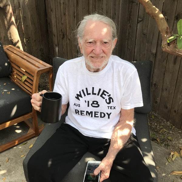 Willie Nelson sitting outdoors holding a mug and phone, wearing a white t-shirt with Willie's Remedy on it.