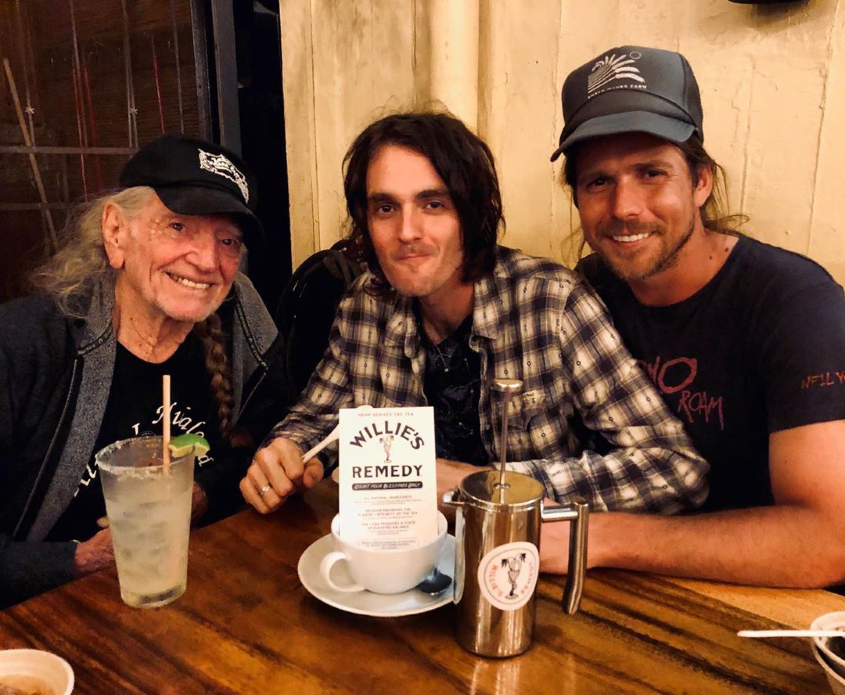 Three men sitting at a table with drinks and a sign for 'Willie's Remedy' in a casual setting.