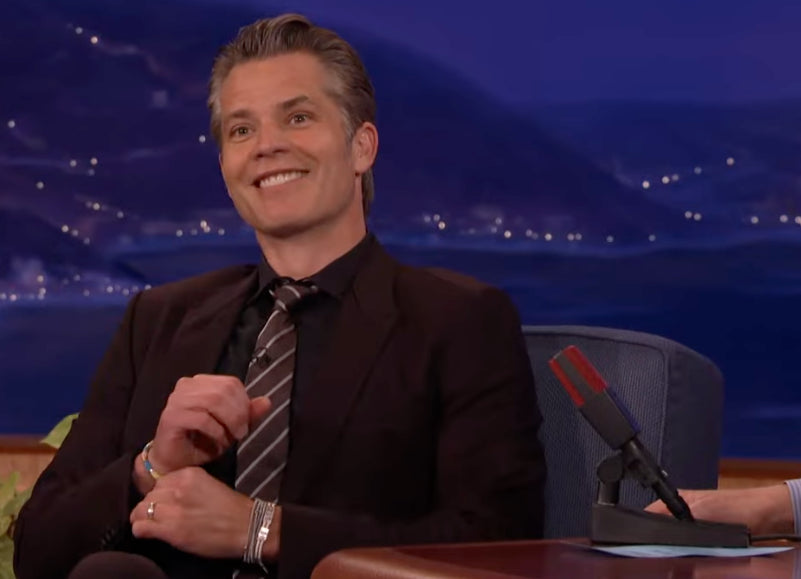 Timothy Olyphant in a suit sitting on a talk show set with a blue background