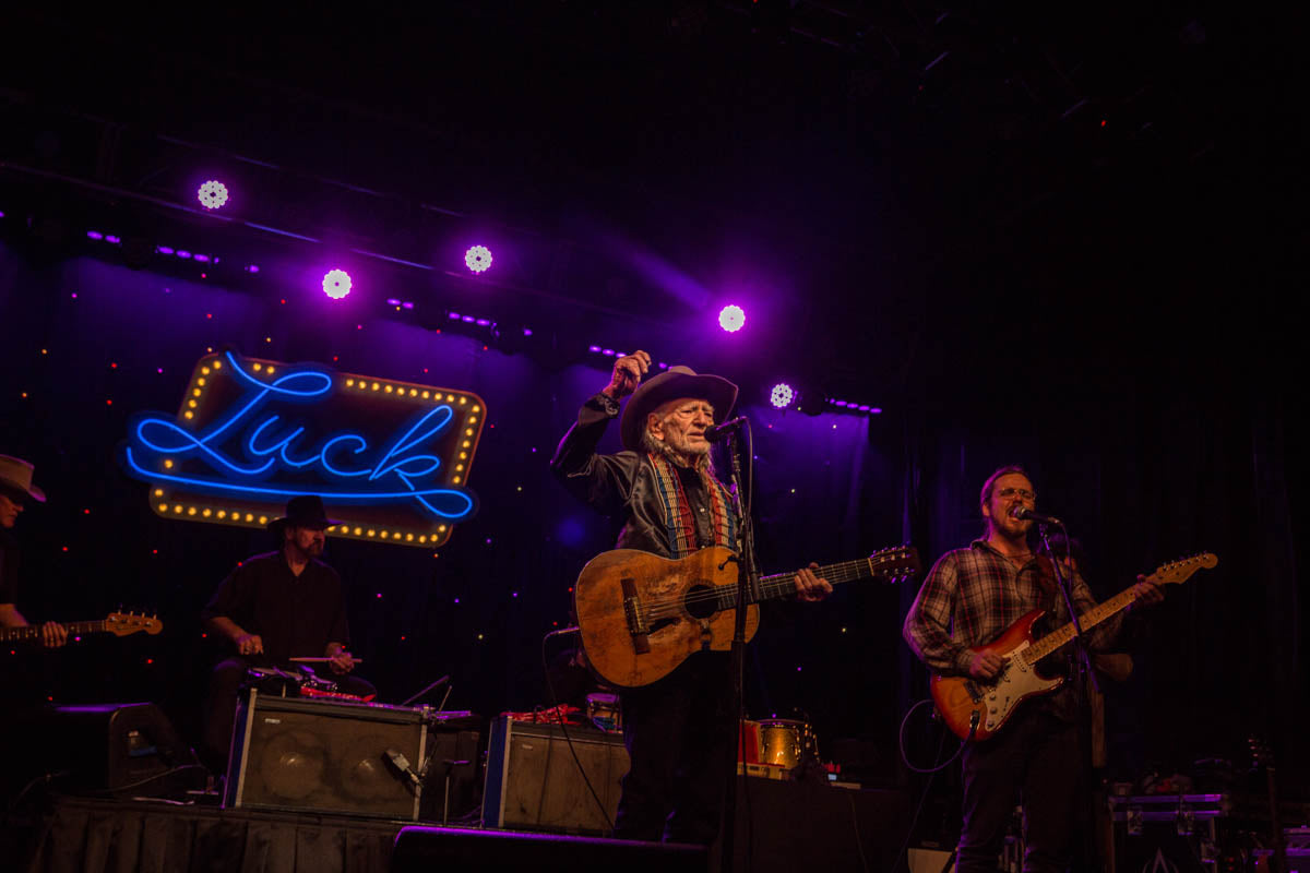 Willie Nelson & band performing on stage with a neon sign in the background that says Luck