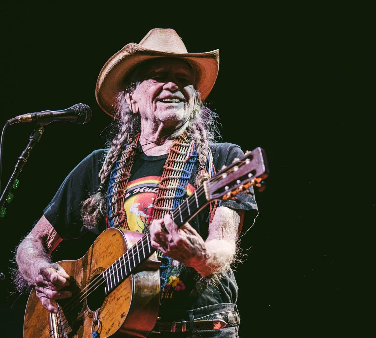 Willie Nelson playing guitar and singing into a microphone with a dark background