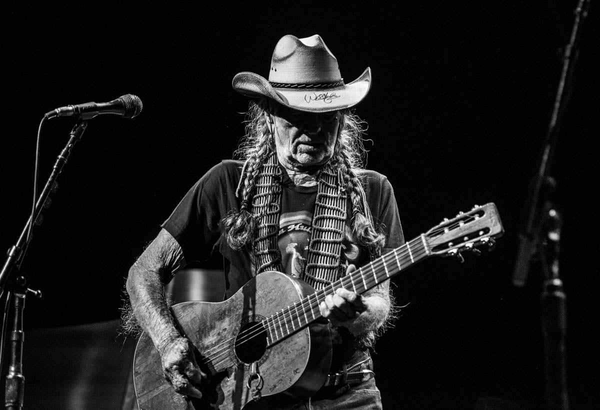 Willie Nelson playing guitar on stage with microphone and dark background
