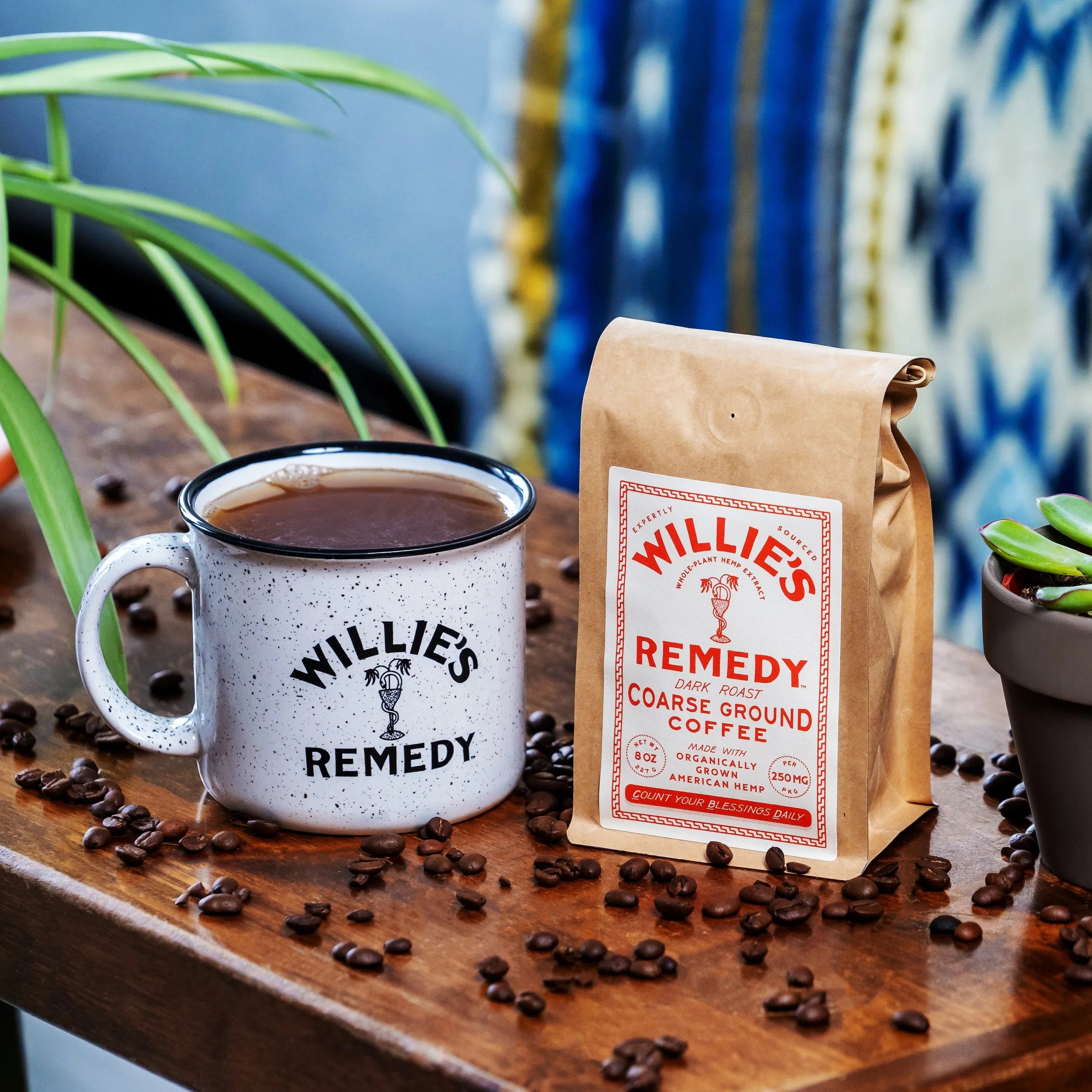 White mug with 'Willie's Remedy' branding next to a brown paper bag of coffee on a wooden surface with scattered coffee beans.